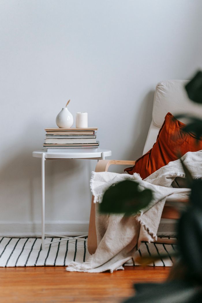 Vases and stack of books placed on small table near comfortable armchair with cushion and plaid in cozy apartment