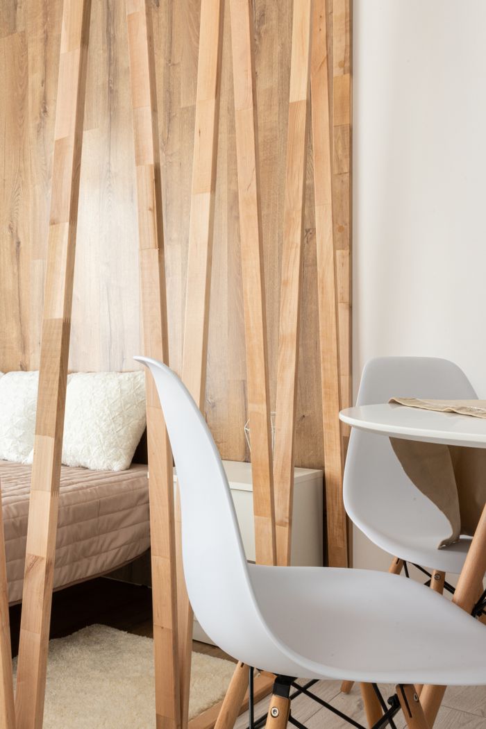 Dining room and bedroom interior with table and chairs against wooden partition and bed with cushions at home in daytime